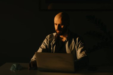 Sakallı, beyaz bir adam karantina sırasında koronavirüsü yaymamak için dizüstü bilgisayarıyla uzaktan çalışıyor. COVID-19 salgını sırasında akşamları evden çalışan bir işadamı.
