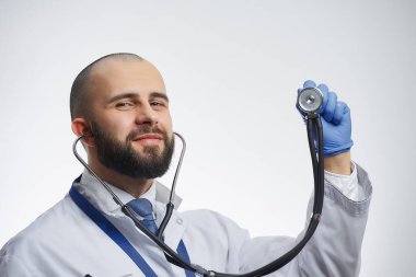 Elinde steteskopla poz veren mutlu bir doktor mavi bir tıp eldiveniyle. Beyaz önlüklü ve mavi kravatlı sakallı bir doktor..