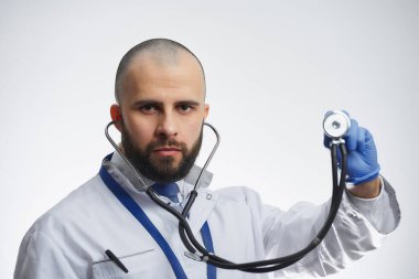 Ciddi bir doktor elinde steteskop tutan mavi bir tıbbi eldiven. Beyaz önlüklü ve mavi kravatlı sakallı bir doktor..