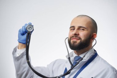 Elinde steteskopla poz veren mutlu bir doktor mavi bir tıp eldiveniyle. Beyaz önlüklü ve mavi kravatlı sakallı bir doktor..