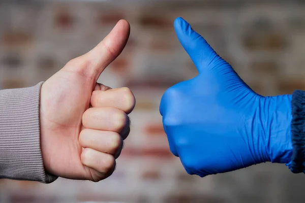 Two thumbs up. A woman thumbs up in a disposable medical glove to a man who gives her thumbs up too with a naked hand. Two friends meet near a brick wall.