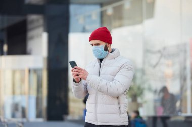 Yayılmış koronavirüsten (COVID-19) kaçınmak için tıbbi yüz maskesi takan beyaz bir adam. Salgın yüzünden yüzünde cerrahi maske olan bir adam şehrin merkezinde akıllı telefon tutuyor..