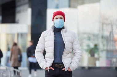 Yayılan koronavirüsten (COVID-19) kaçınmak için tıbbi yüz maskesi takan bir erkek. Yüzünde maske olan bir adam. Salgın yüzünden şehrin göbeğindeki kot pantolonun cebinde el ele tutuşuyor..