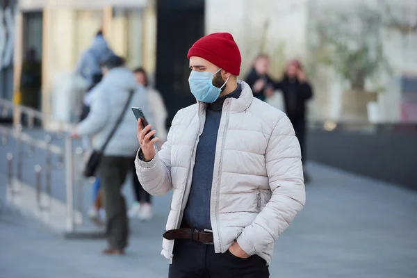 A caucasian man wearing a medical face mask to avoid the spread coronavirus (COVID-19). A guy with a surgical mask on the face holding a smartphone in the center of the town.