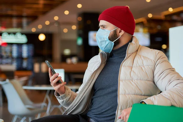 Koronavirüsten (COVID-19) korunmak için tıbbi yüz maskesi takan sakallı beyaz bir adam. Yüzünde cerrahi maske olan bir adam alışveriş merkezinde akıllı bir telefonla iş yapıyor.