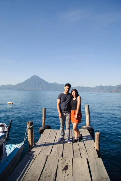 Pareja viajera parada en el muelle del lago Atitlán sonriendo a la cámara - amanecer en el lago Guatemala - Pareja hispana de aventura en destino turístico de América Latina — Foto de Stock