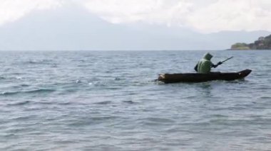 Göl, göl ve mavi gökyüzü manzarasının ortasında küçük bir kayıktaki adam kıyıya gelen ahşap motorlu tekne, Guatemala 'daki volkanlar ve dağlarla çevrili.