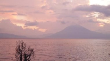 Guatemala 'daki temiz atitlan gölünde gün batımı. Gün batımında gölün ortasında küçük bir tekne. Gökyüzünde mor renkler olan bulutlar, volkanlar ve dağlar arasında.