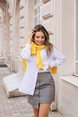 Beyaz tişörtlü genç kız açık bej bir binanın önünden geçiyor.