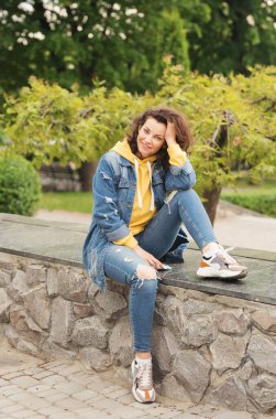 Kıvırcık saçlı genç bir kadın şehir parkında bir kaldırımda oturuyor. Üzerinde kot pantolon ve elinde telefonla gülümseyen parlak sarı bir kapüşonlu var.