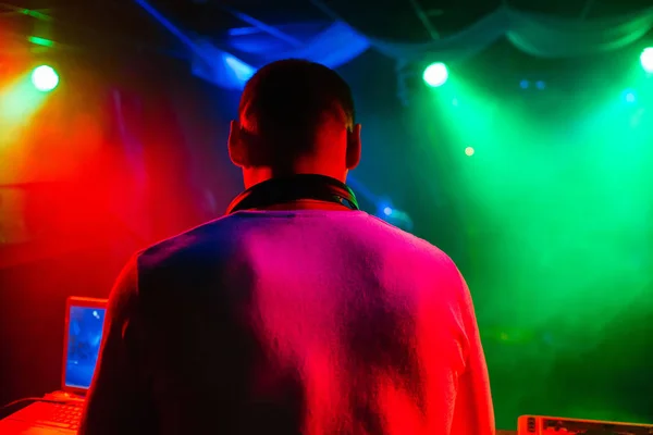 DJ desde atrás en discoteca con auriculares mezclan música en discoteca con fondo colorido —  Fotos de Stock