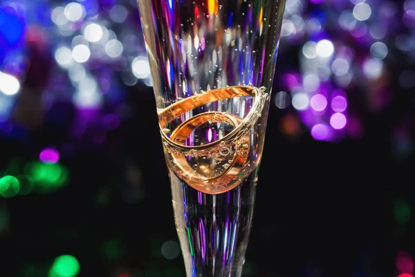 Золотые обручальные кольца в бокале шампанского с пузырьками на цветном размытом фоне — стоковое фото