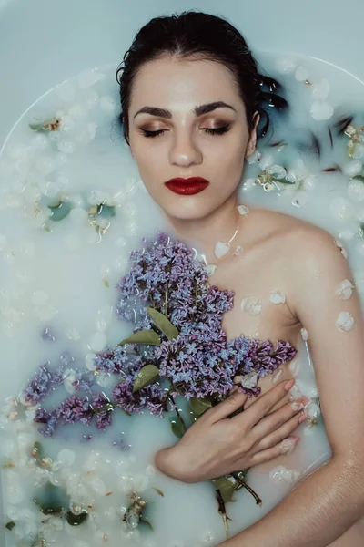 Retrato de beleza de uma bela menina desfrutando e relaxando no banho com leite e flores de maçã com um buquê de lilases — Fotografia de Stock