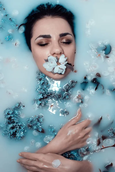 Rosto de uma jovem com uma flor na boca e um buquê de lilases relaxando e desfrutando no banho com leite azul — Fotografia de Stock