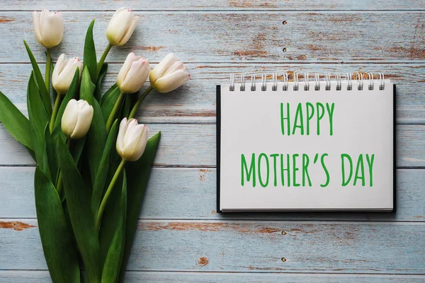 Boeket witte bloemen van tulpen op de achtergrond van blauwe planken met een notitieboekje met belettering Happy Mothers Day — Stockfoto