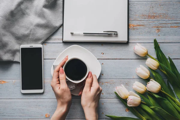 Strauß weißer Tulpen mit einer Tasse Kaffee in Frauenhand, einem Smartphone und einem leeren Notizbuch für den Text — Stockfoto