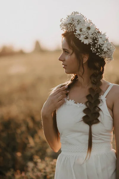Portret van aantrekkelijk meisje met bloem krans en vlechten in witte jurk in zomer veld bij zonsondergang — Stockfoto