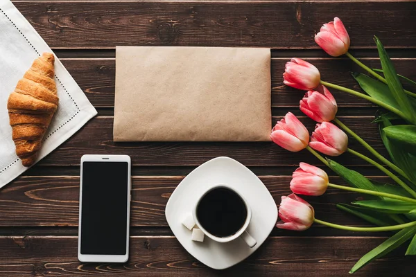 Tasse Kaffee mit einem Croissant zum Frühstück auf dem Tisch mit einem Strauß rosa Tulpen und einem Smartphone dekoriert — Stockfoto