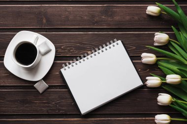 Bir fincan kahve ve boş bir not defteri ile ahşap bir masa üzerinde beyaz laleler