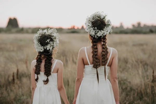Mamma e figlia dorsi in abiti bianchi con trecce e ghirlande floreali in stile boho in estate — Foto Stock