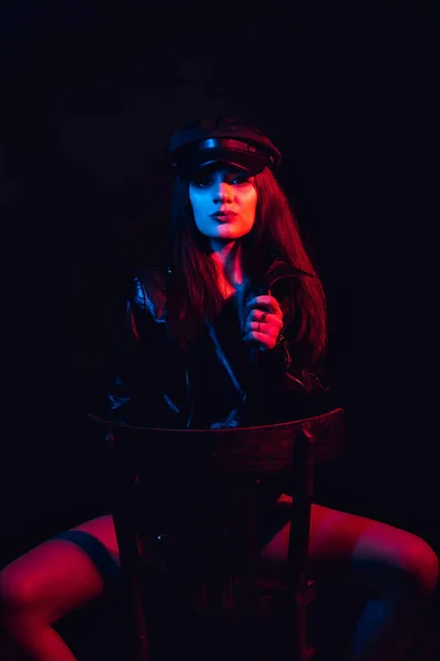 Retrato elegante de uma jovem menina sexy em uma jaqueta de couro e boné com uma luz de néon — Fotografia de Stock