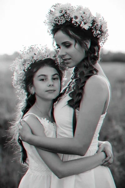 Mãe e filha abraçando juntos em vestidos brancos com tranças e coroas florais no estilo boho no verão — Fotografia de Stock