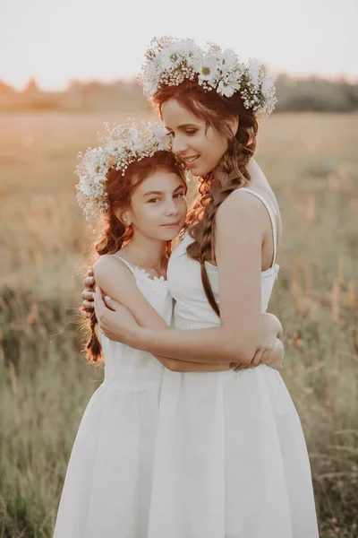 Mãe e filha abraçando juntos em vestidos brancos com tranças e coroas florais no estilo boho no verão — Fotografia de Stock