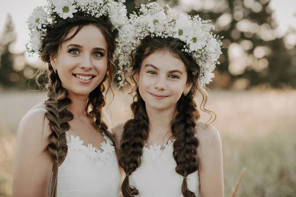 Felice mamma e figlia in abiti bianchi con ghirlande floreali e trecce in stile boho in estate in un campo — Foto Stock