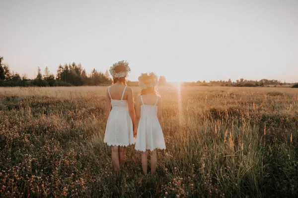 Mor och dotter stå tillbaka i sommaren vid solnedgången i fältet i vita klänningar och blommor kransar — Stockfoto