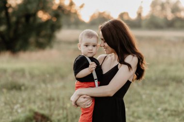 young happy mom holds her son boy in her arms in a meadow in summer at sunset