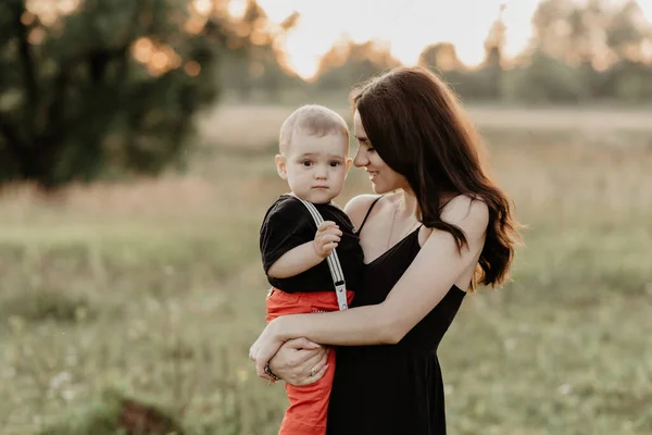 Joven feliz mamá sostiene a su hijo niño en sus brazos en un prado en verano al atardecer —  Fotos de Stock