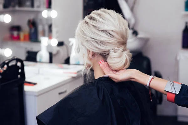Парикмахер превращает волосы в булочку на голове блондинки с длинными волосами — стоковое фото
