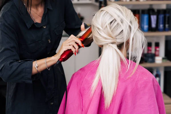 Mãos de um cabeleireiro com um ferro fazer o cabelo de uma menina loira — Fotografia de Stock