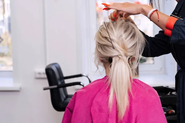 Mãos de um cabeleireiro profissional fazer um penteado baixo bun para uma menina loira — Fotografia de Stock