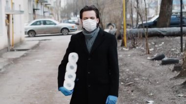 Koronavirüs salgınına karşı korunmak için eldiven ve tıbbi maske takan adam tuvalet kağıdı rulolarıyla korunuyor.