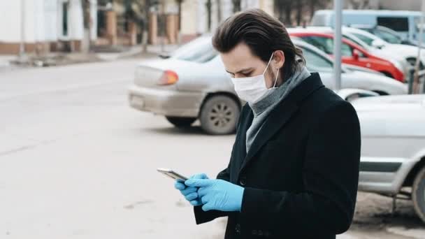 Hombre blanco caucásico que usa una máscara médica respiratoria y guantes para protegerse contra el coronavirus COVID-19 utiliza un teléfono inteligente — Vídeo de stock