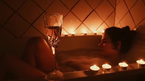 Бокал шампанского со свечами в ванной комнате на фоне молодой расслабляющей девушки, принимающей ванну с пеной — стоковое видео