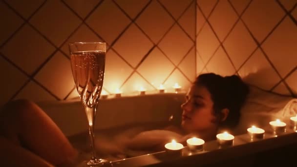 Бокал шампанского со свечами в ванной комнате на фоне молодой расслабляющей девушки, принимающей ванну с пеной — стоковое видео