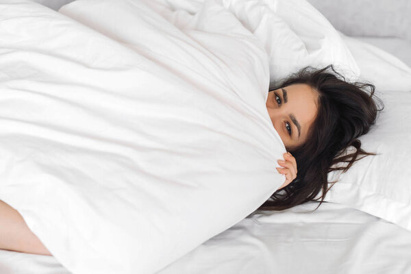 young beautiful Caucasian girl looks out from under the blanket on a white bed