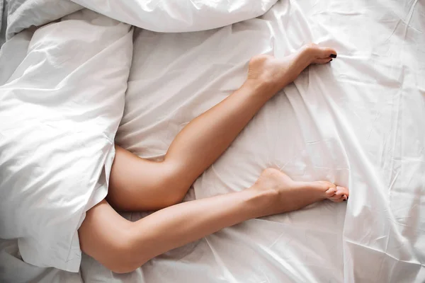 Piernas delgadas de una joven sobresalen de debajo de la manta en una sábana blanca — Foto de Stock