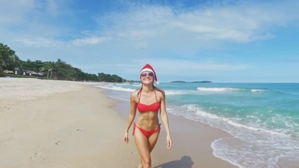 Bela mulher de Natal caucasiana em chapéu de santa e piadas de biquíni vermelho e relaxante na praia com areia branca. Ano Novo na selva vista para o mar e luz solar. Férias em trópicos . — Vídeo de Stock