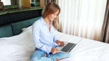 Yatak odasında dizüstü bilgisayarda çalışan güzel beyaz kadın. MacBook kullanarak ve gülümseyen genç tatlı kız. Güzel mutlu teen yatakta oturma ve sınavları için hazırlanıyor. Güneşli sabah.