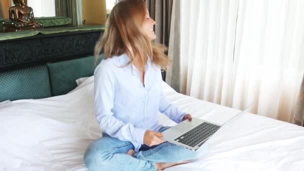 美丽的白种女人在卧室的笔记本电脑上工作。年轻可爱的女孩使用 macbook 和微笑。好快乐的少年坐在床上，准备考试。阳光明媚的早晨. — 图库视频影像