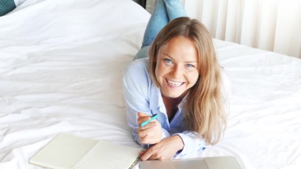 Hermosa mujer caucásica trabajando en el ordenador portátil en el dormitorio. Joven linda chica hablando por teléfono y sonriendo. Niza adolescente feliz se encuentra en la cama y se prepara para los exámenes. Mañana soleada . — Vídeo de stock