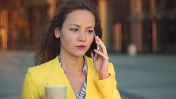 Mooi meisje in een gele jas in de buurt van office blijven en glimlachend op het balkon met de koffie. Glimlachende zakenvrouw met een kopje thee op het business center. Fel zonlicht in de ochtend. — Stockvideo