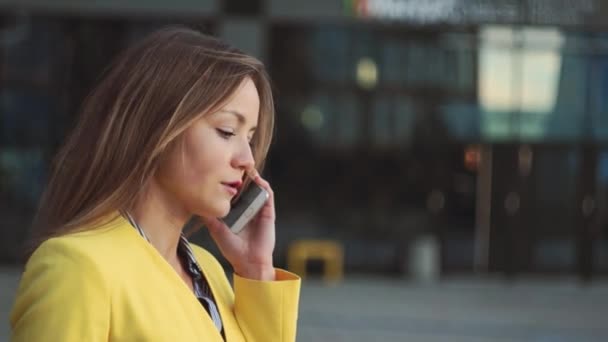 Portret van jonge mooi meisje in een gele jas in de buurt van office drinken koffie praten via de telefoon. Glimlachende zakenvrouw met een kopje thee op het business center. — Stockvideo