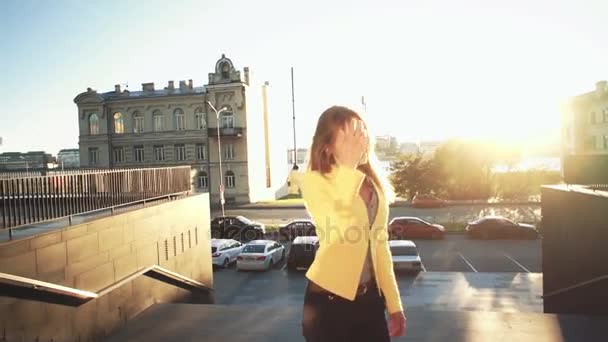 Movimento lento. Menina bonita nova em uma jaqueta amarela subindo as escadas e falando pelo telefone. Mulher de negócios caminhando para trabalhar no centro de negócios. Luz solar brilhante pela manhã . — Vídeo de Stock