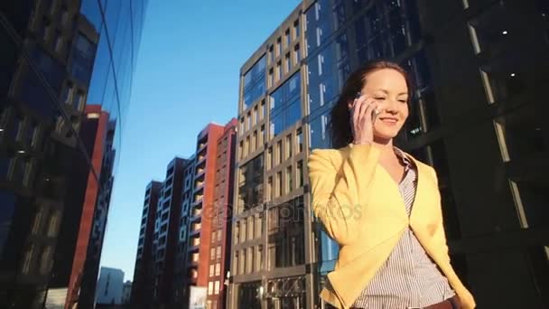 Bajo ángulo ver video de Joven hermosa chica en una chaqueta amarilla cerca de la oficina con el teléfono. Mujer de negocios hablando por teléfono inteligente en el centro de negocios. Luz del sol brillante en la mañana . — Vídeos de Stock