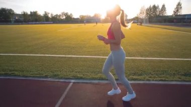 Koşu ve atletik yolda stadyum üzerinde güneş doğarken çalışan güzel bir genç kadın egzersiz. Sinema tarzı video.