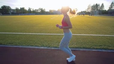 Ağır çekim. Koşu ve atletik yolda stadyum üzerinde güneş doğarken çalışan güzel bir genç kadın egzersiz. Sinema tarzı video.
